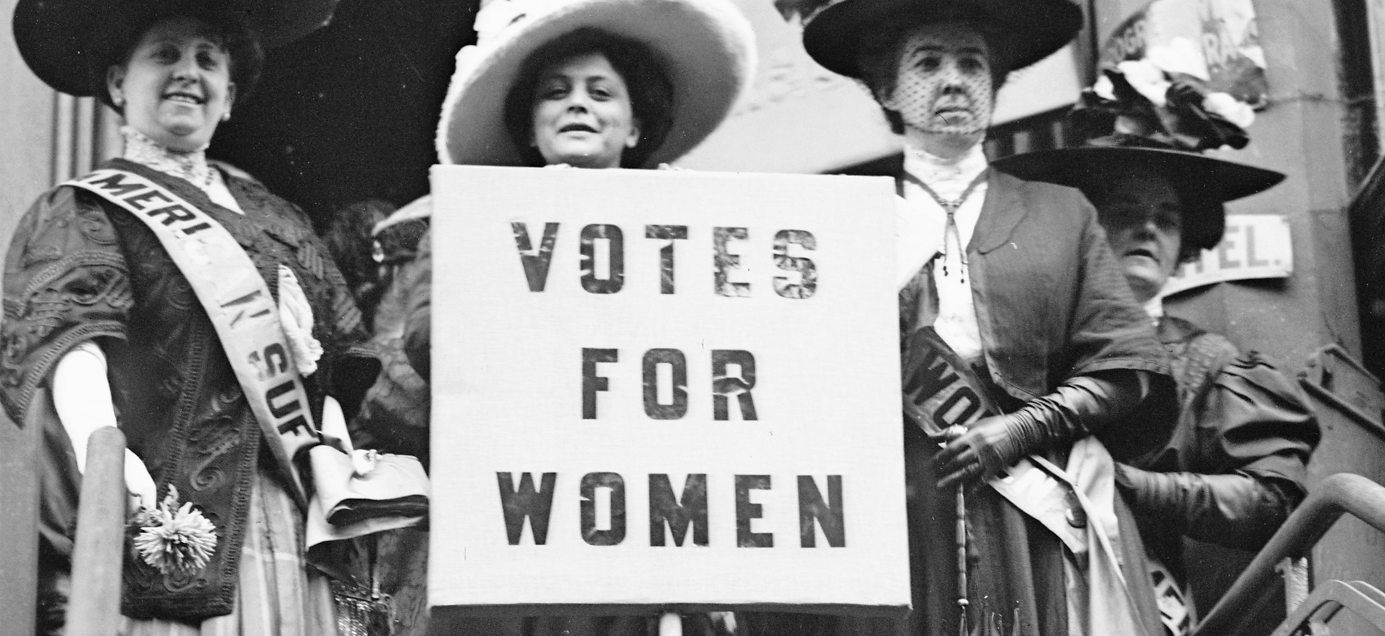 Historical, back and white photo of the Women's Suffrage movement with three women holding a sign that states Votes for Women.