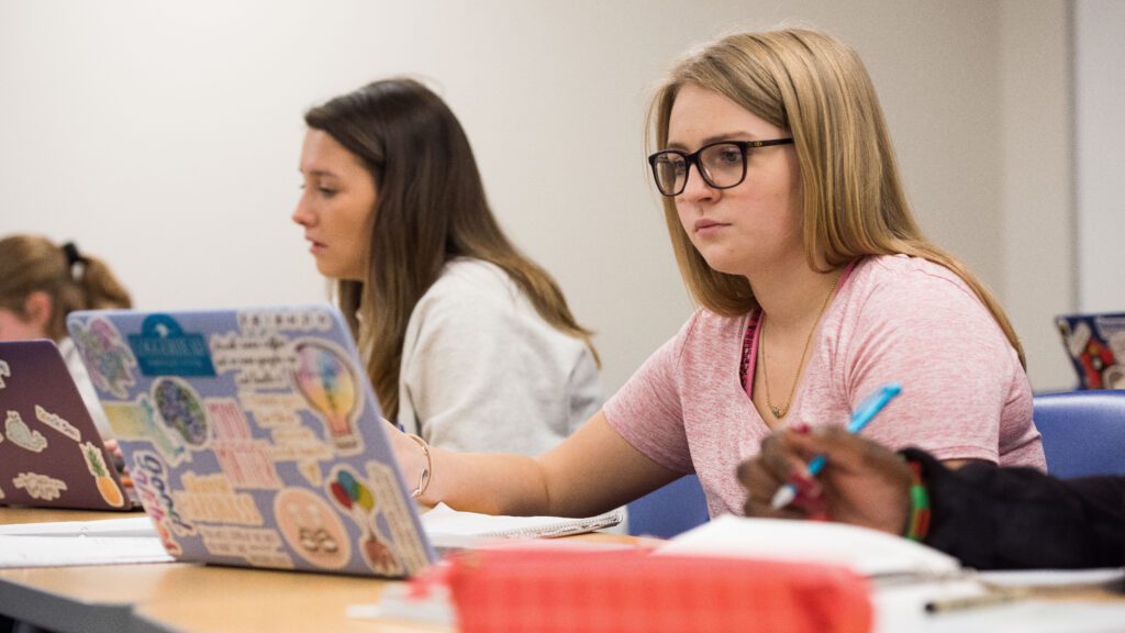 A female student in a pink shirt and glasses using her laptop in class.