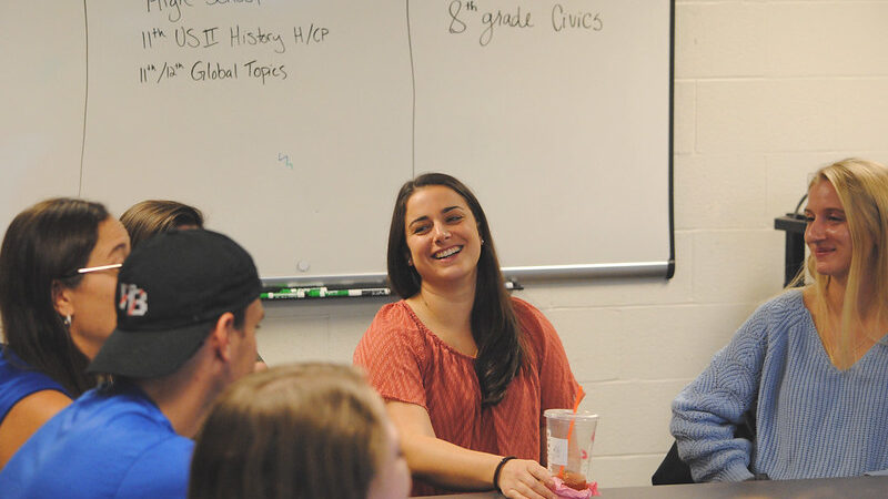 Students gathered around a circle table with a whiteboard in the background.